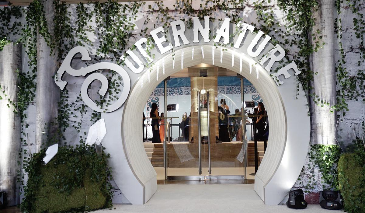 A stylish arched entrance decorated with ivy and greenery displays the word SUPERNATURE above glass doors, with people visible inside through the entrance.