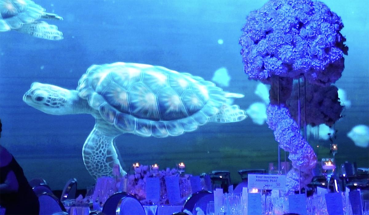 A large projection of a sea turtle swims across a blue-lit wall above a decorated dining area with floral centerpieces, candles, and elegant place settings.
