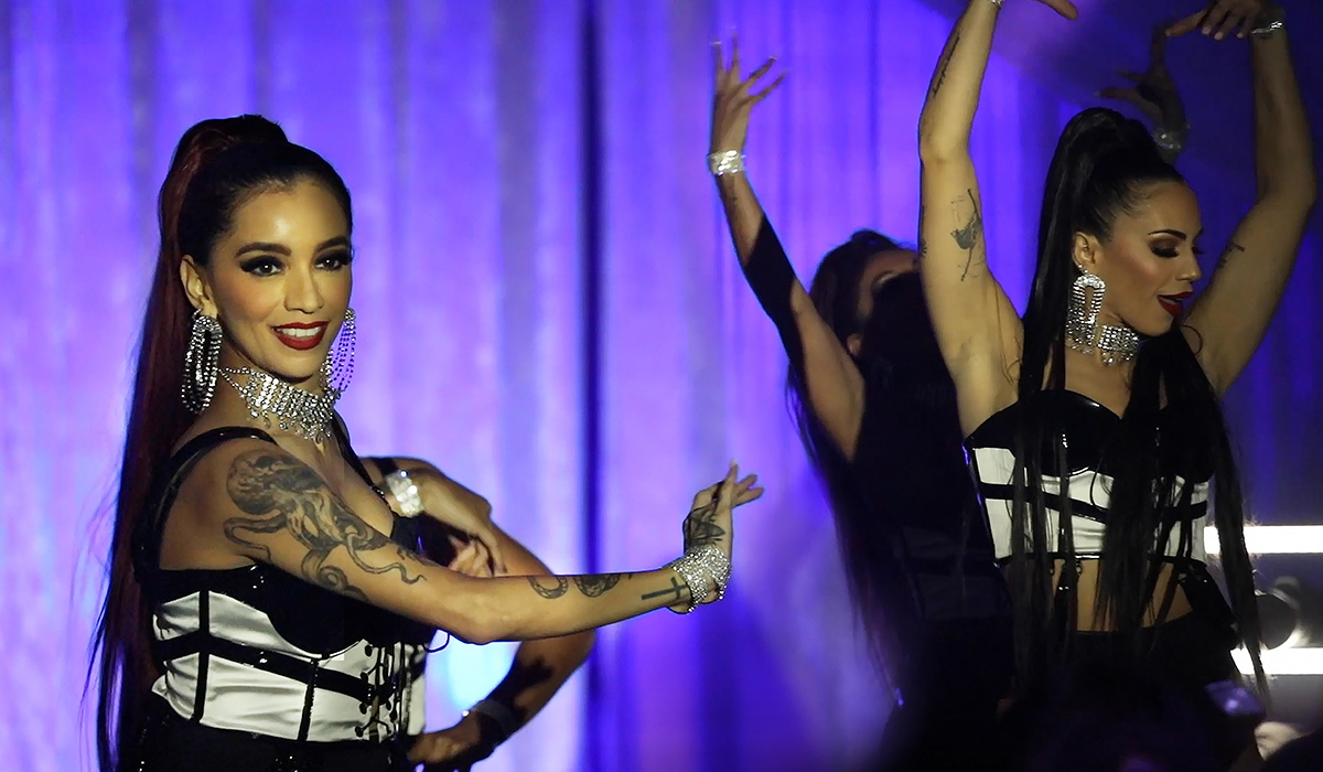 Two women with long dark hair, dramatic makeup, large earrings, and glittering jewelry perform a choreographed dance on stage, wearing black and white outfits, against a purple-lit backdrop.