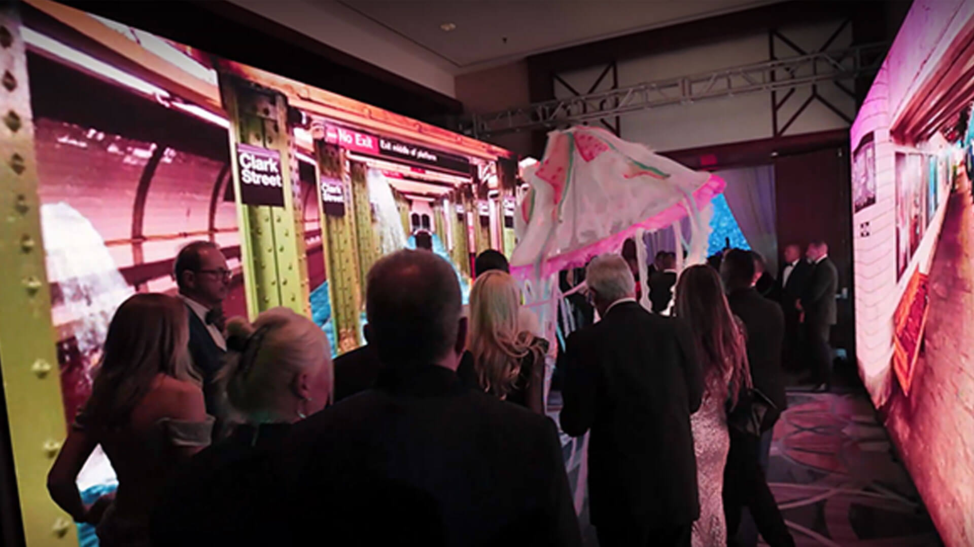 A group of people in formal attire stand near a large, brightly lit display featuring a subway train. Some are holding a decorative umbrella, creating a festive atmosphere in the indoor event space.