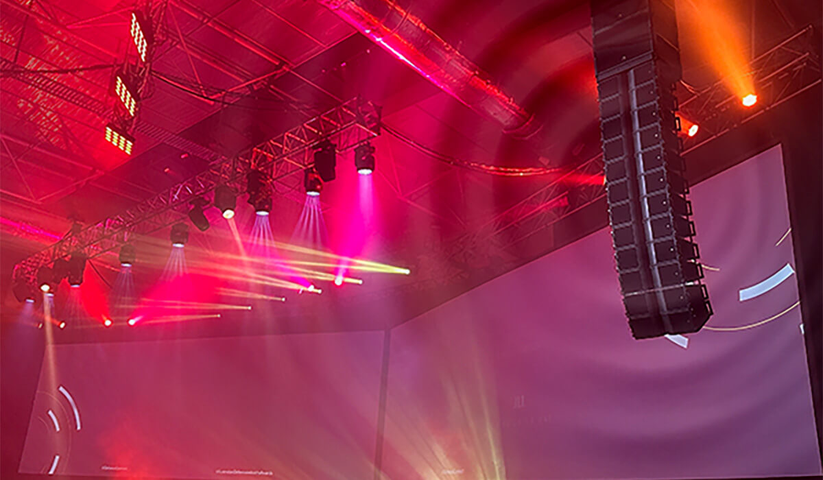 A concert or event stage with bright pink, red, and yellow spotlights shining through fog, large hanging speakers, and big projection screens in the background.