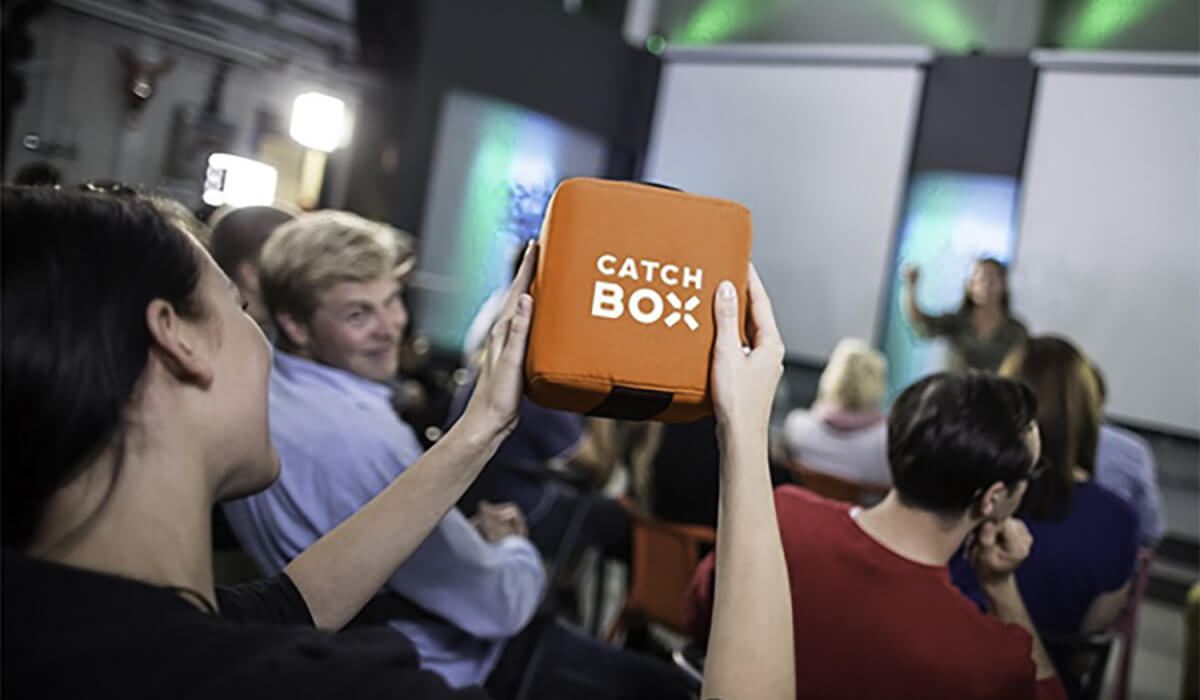 A person holds up an orange cube-shaped microphone labeled Catch Box during an event, while others sit and face a speaker at the front of the room.