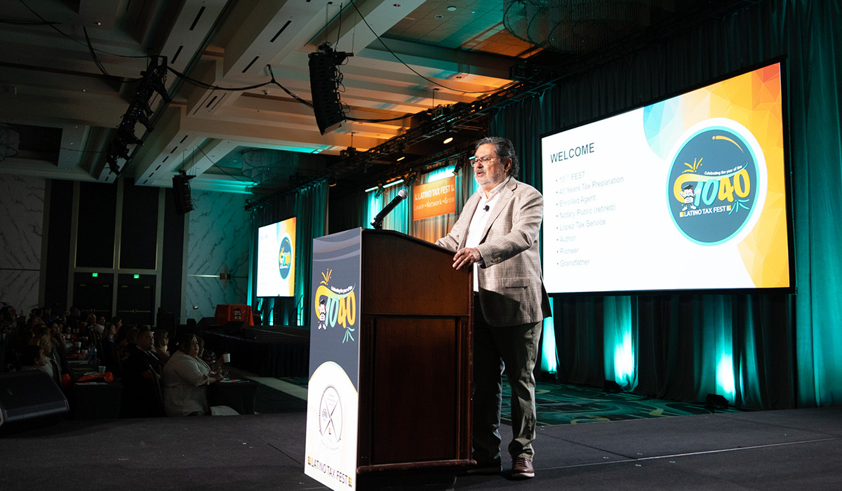 A man in a light-colored suit stands at a podium on a stage, addressing an audience. Behind him, large screens display a WELCOME message and a colorful logo. The room is dimly lit with blue-green accent lighting.