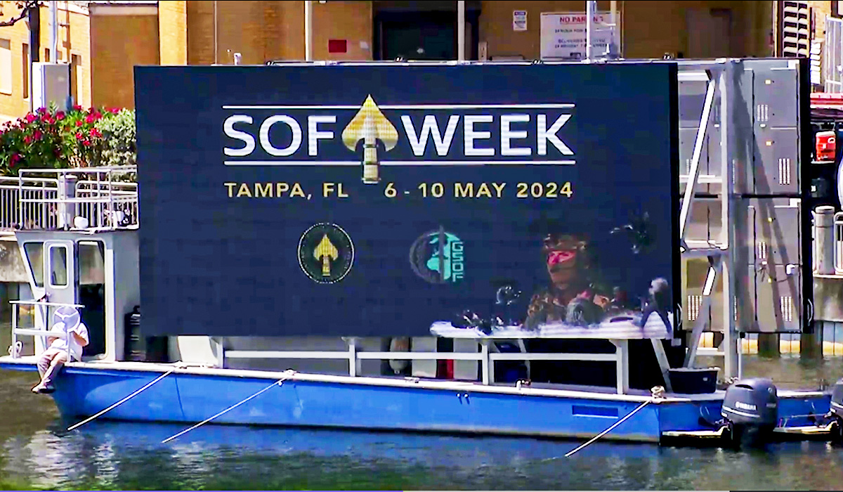 A large digital sign on a docked boat displays SOF WEEK, Tampa, FL, 6-10 May 2024 with logos and images of soldiers. A person sits at the front of the blue boat on a sunny day.
