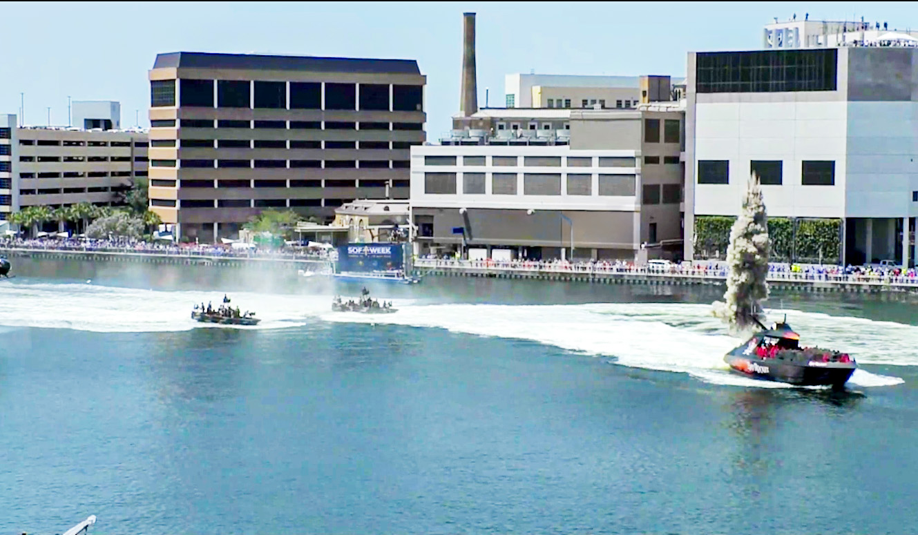 Two boats speed through a river in front of city buildings, creating large wakes. A tall splash or water plume rises near the shore, and a crowd watches from behind a barrier along the waterfront.