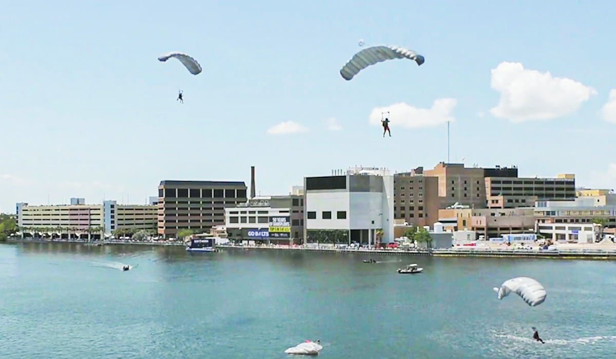 Four skydivers with open parachutes descend over a calm body of water near a city shoreline with several modern buildings and a partly cloudy sky. Small boats and people are visible on the water below.