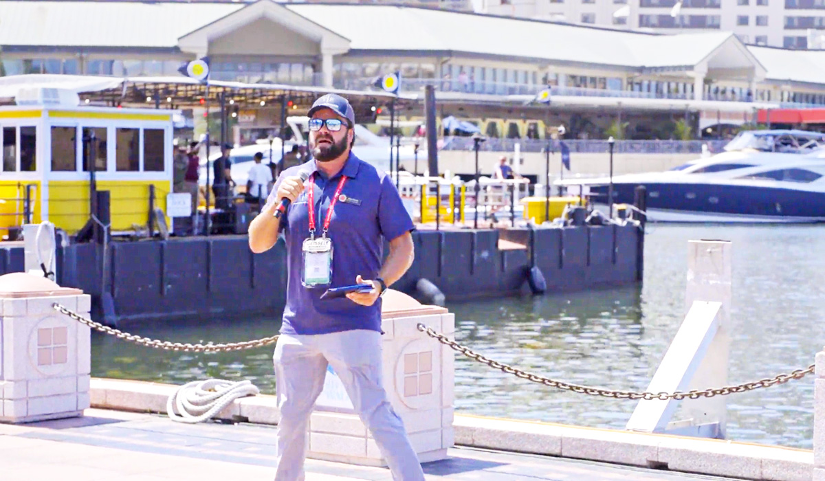 A man in sunglasses and a cap holds a microphone and tablet while standing near a waterfront marina with boats and a building in the background on a sunny day.