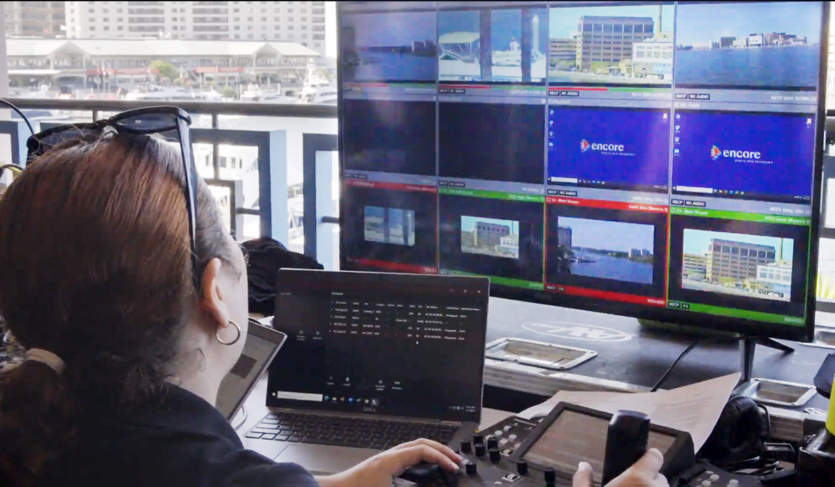 A person operates video production equipment, including a laptop and control panel, while monitoring multiple live video feeds displayed on large screens with waterfront buildings visible in some camera views.