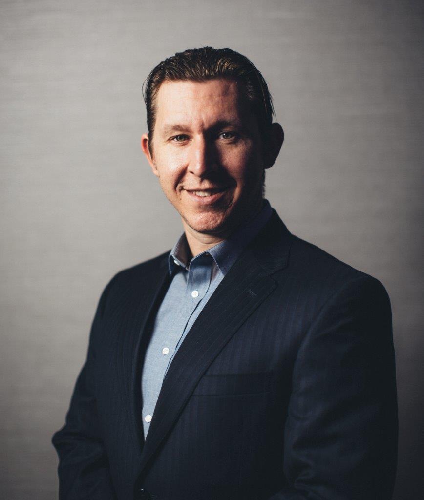 A man in a dark suit and blue shirt smiles at the camera against a plain, softly lit background.