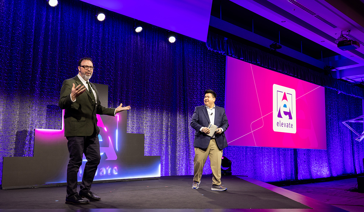 Two men stand on a brightly lit stage with a large screen displaying the word elevate and a stylized “A” logo behind them. The background is purple with stage lights shining down.