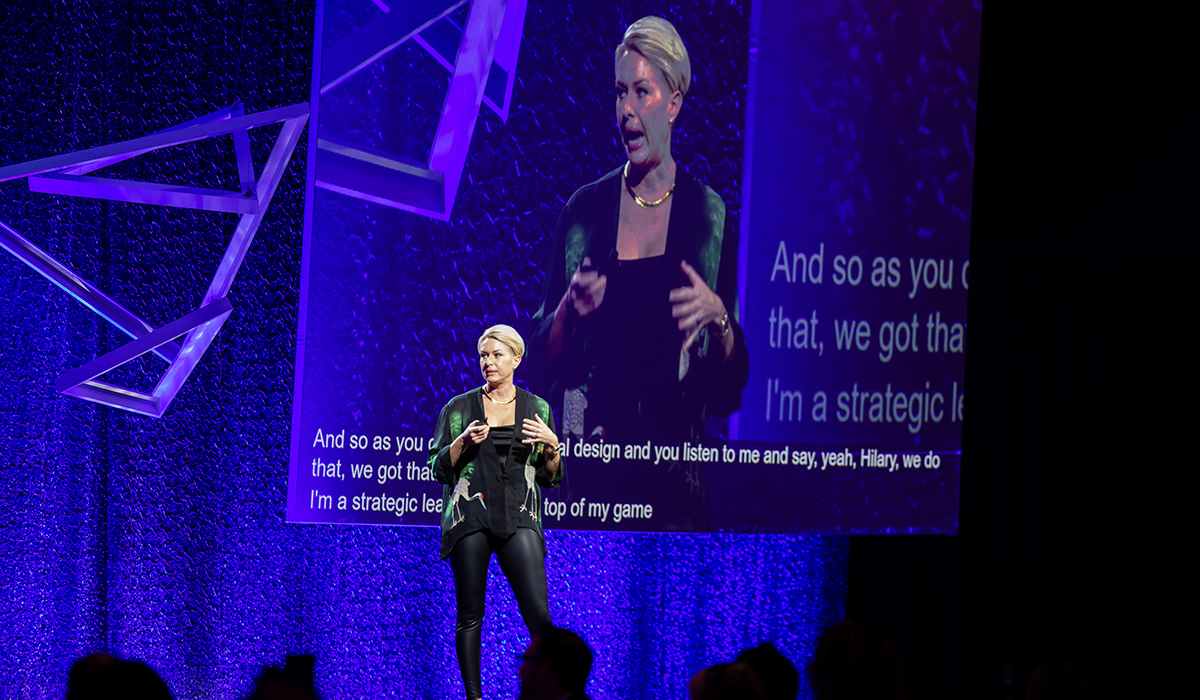 A woman stands on stage giving a presentation with subtitles displayed behind her. She gestures as she speaks, and a large projection of her image is visible on a screen. The background is decorated with abstract geometric shapes and purple lighting.