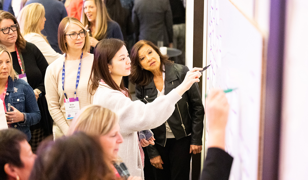 A group of women stands around a whiteboard while one woman writes on it with a marker. The group watches attentively, suggesting a workshop or collaborative meeting.