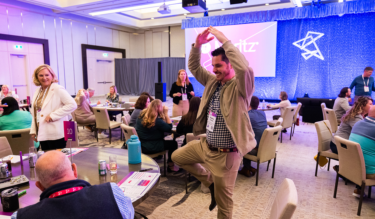 A man in business attire strikes a playful ballet pose in a conference room, smiling as others seated at round tables watch and laugh. Event materials and a stage with a pink sign are visible in the background.
