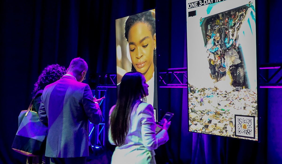 Three people stand in front of two large digital screens; one shows a woman’s face with closed eyes, the other displays an image of debris and a QR code. The scene is lit with colorful lighting.