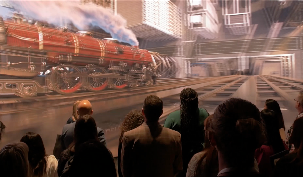A group of people stand indoors facing a large, detailed digital display of a red steam locomotive with blue smoke, shown on railroad tracks in a futuristic setting.