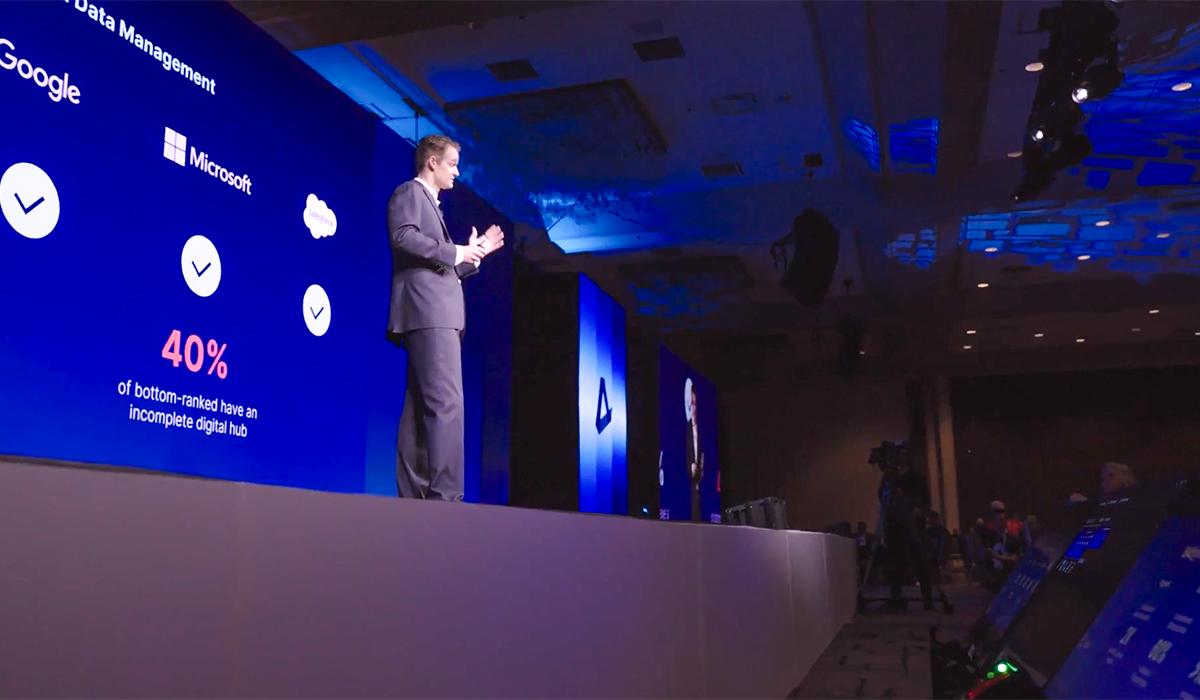 A man in a suit speaks on stage at a conference. Behind him are large blue screens showing logos for Google and Microsoft with text reading “40% of bottom-ranked have an incomplete digital hub.”.