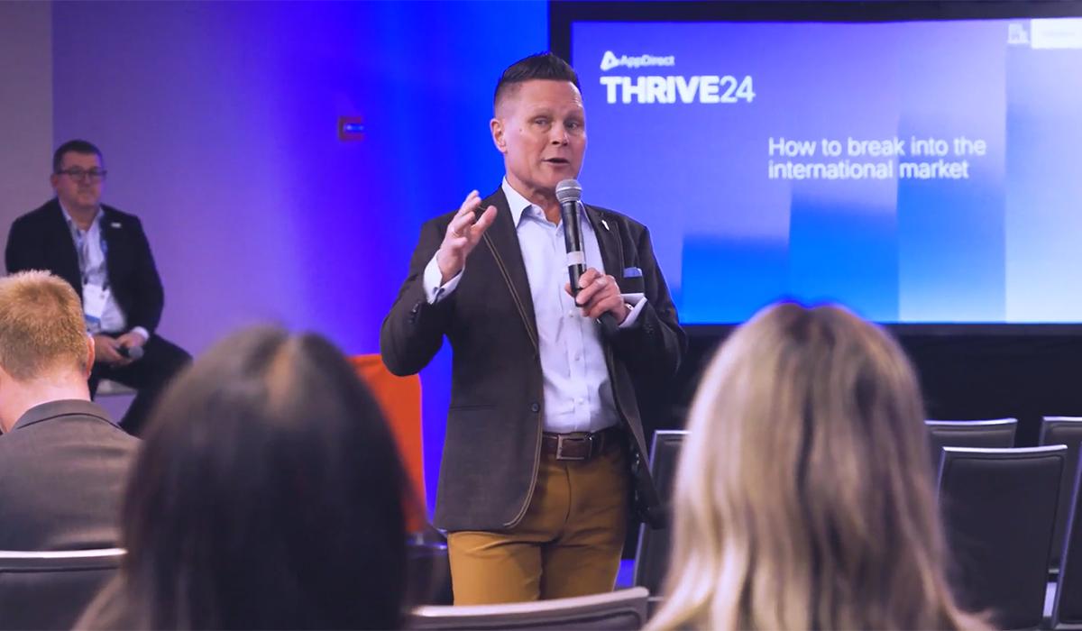 A speaker in a blazer and tan pants holds a microphone and gestures while presenting to an audience. Behind him, a screen displays “THRIVE24: How to break into the international market.”.