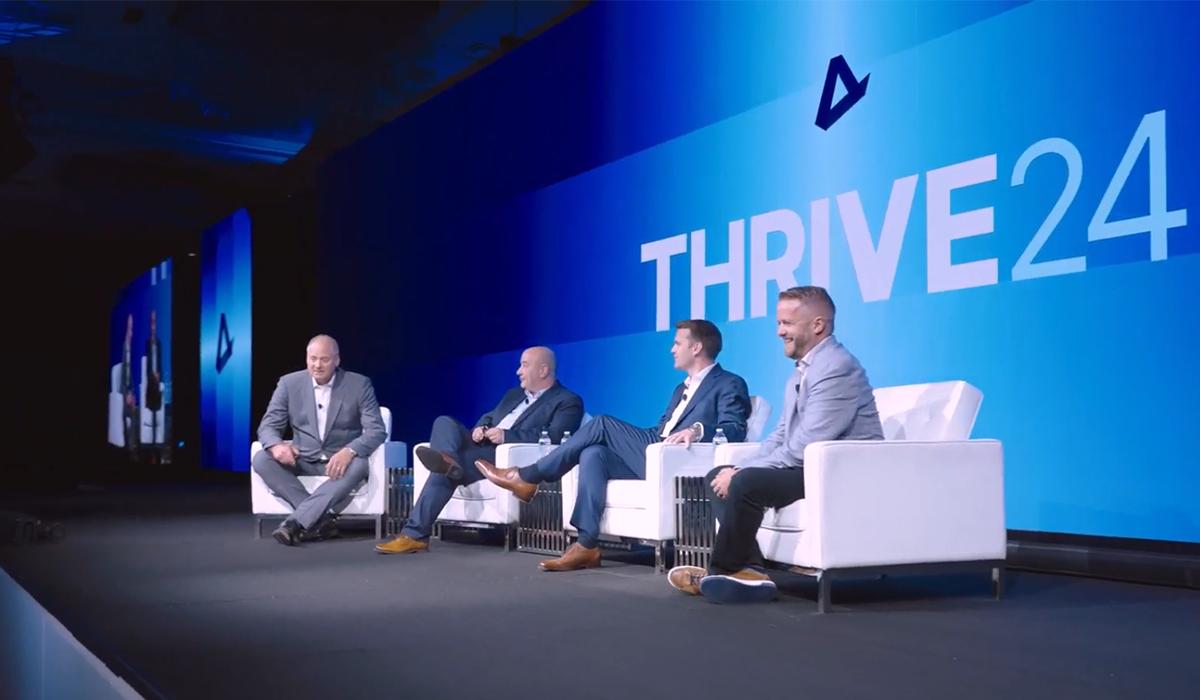 Four men in suits sit on stage in white chairs, engaged in discussion at an event with a large blue backdrop displaying THRIVE24 in bold white letters.
