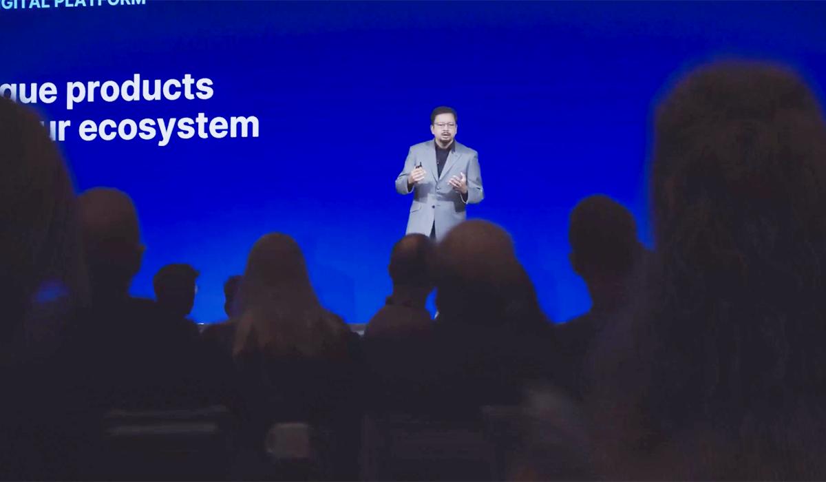 A man in a gray suit speaks on stage in front of a blue screen with white text, addressing an audience seated in the foreground. The text reads, que products ur ecosystem.