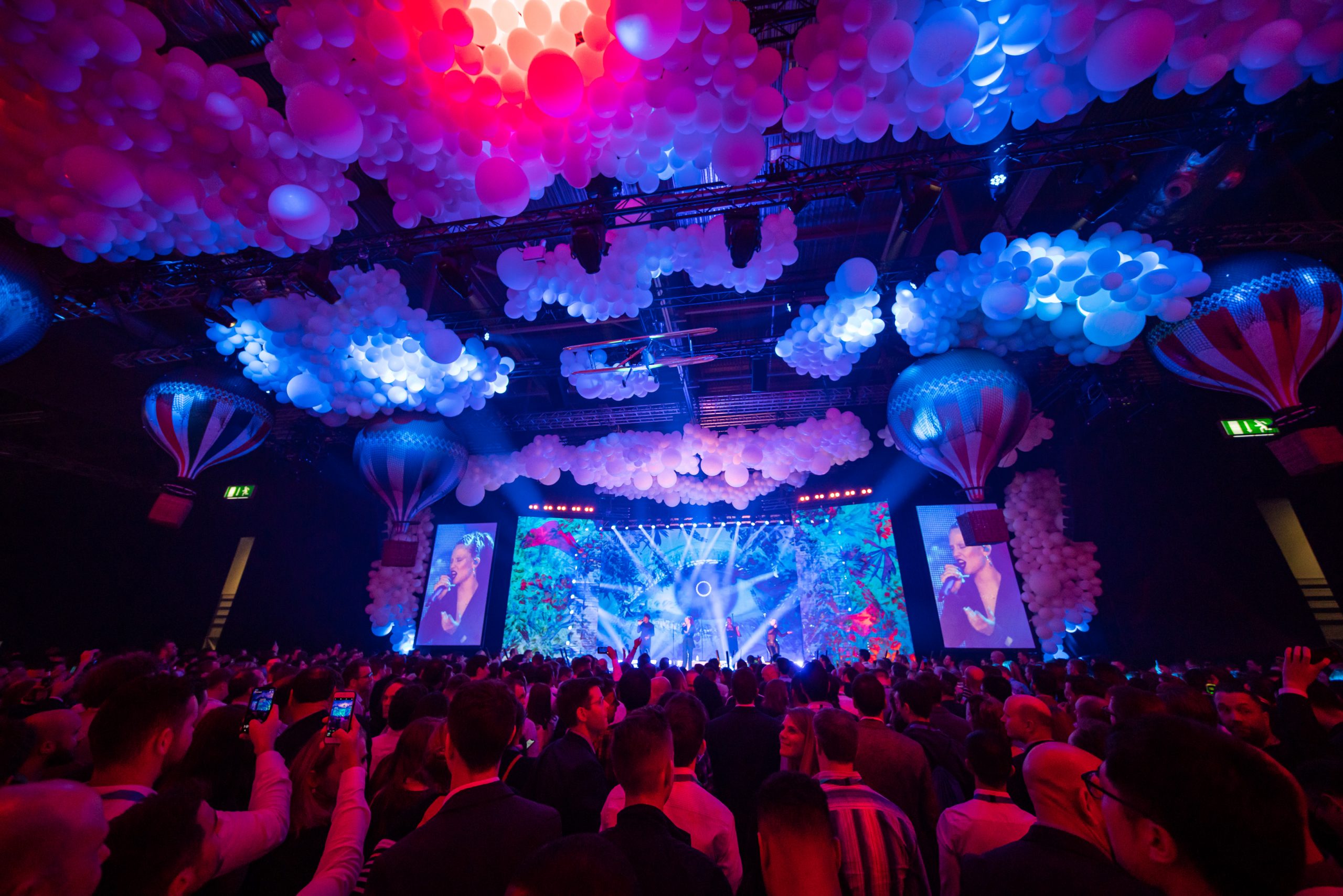 A crowd faces a brightly lit stage with colorful lights and large screens, surrounded by hot air balloon decorations and clusters of white balloons on the ceiling, creating a festive atmosphere.