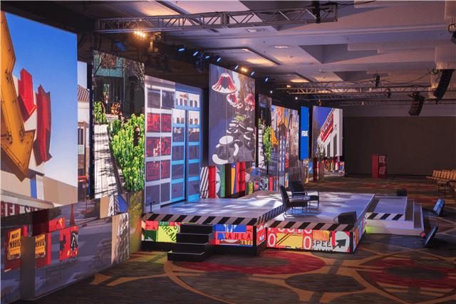 A colorful stage setup with large screens displaying urban and graphic images, a black chair at center stage, and vibrant lighting in a spacious event venue.