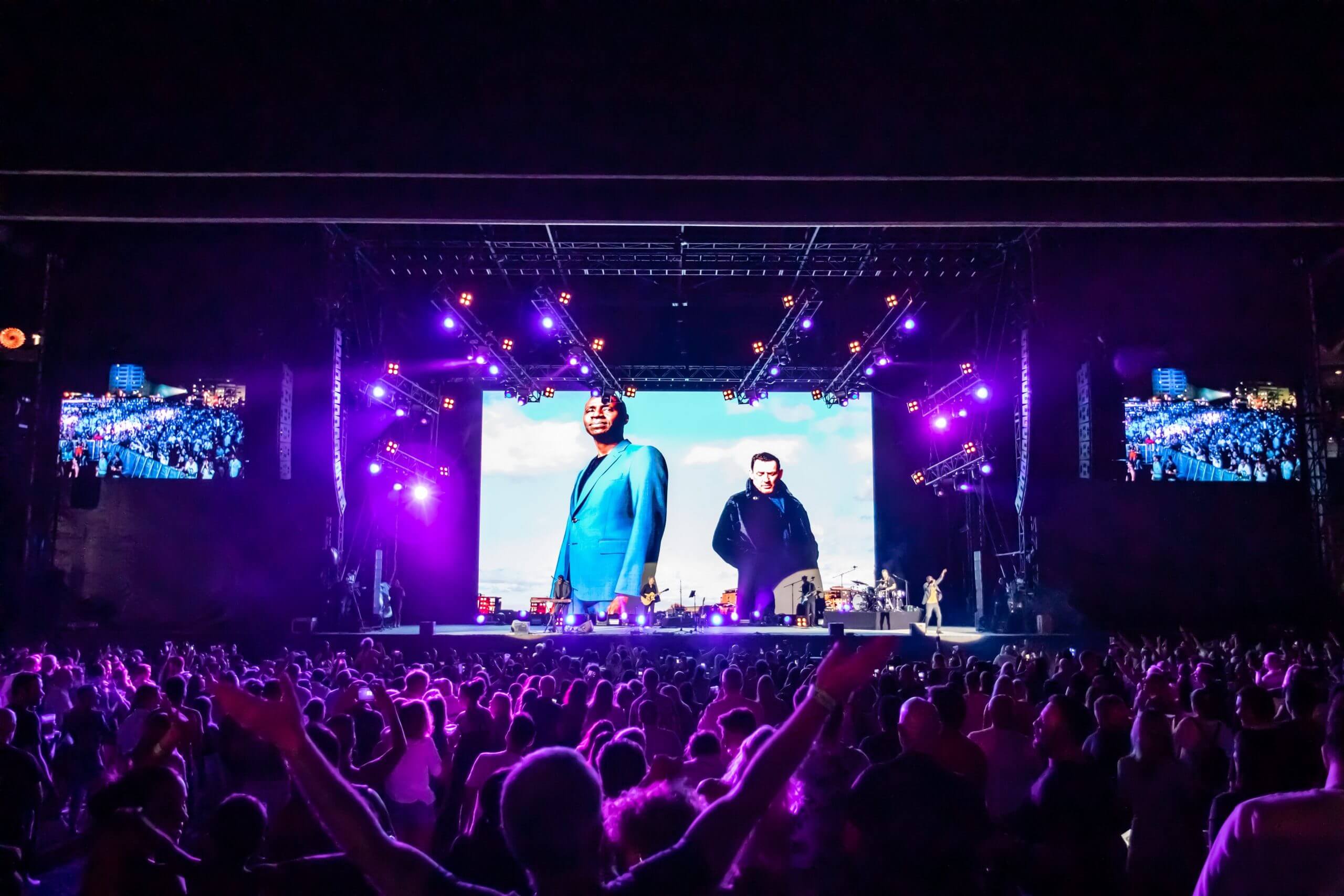 A large crowd at an outdoor concert at night, facing a brightly lit stage with purple lights and a giant screen displaying two men in suits against a sky background.