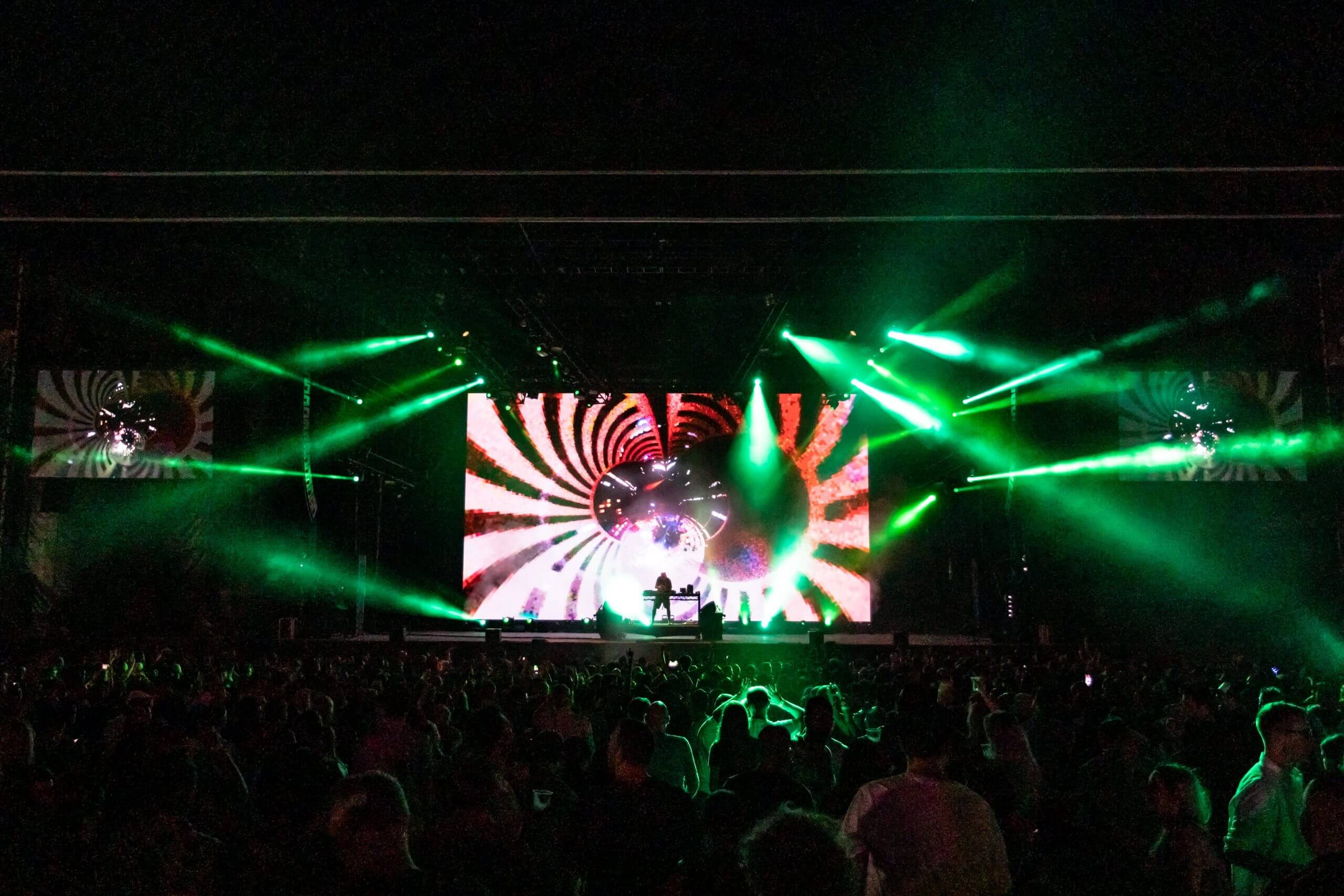 A vibrant concert scene at night with green stage lights, a large psychedelic spiral pattern on the main screen, and silhouettes of a crowd and performers in front of the stage.