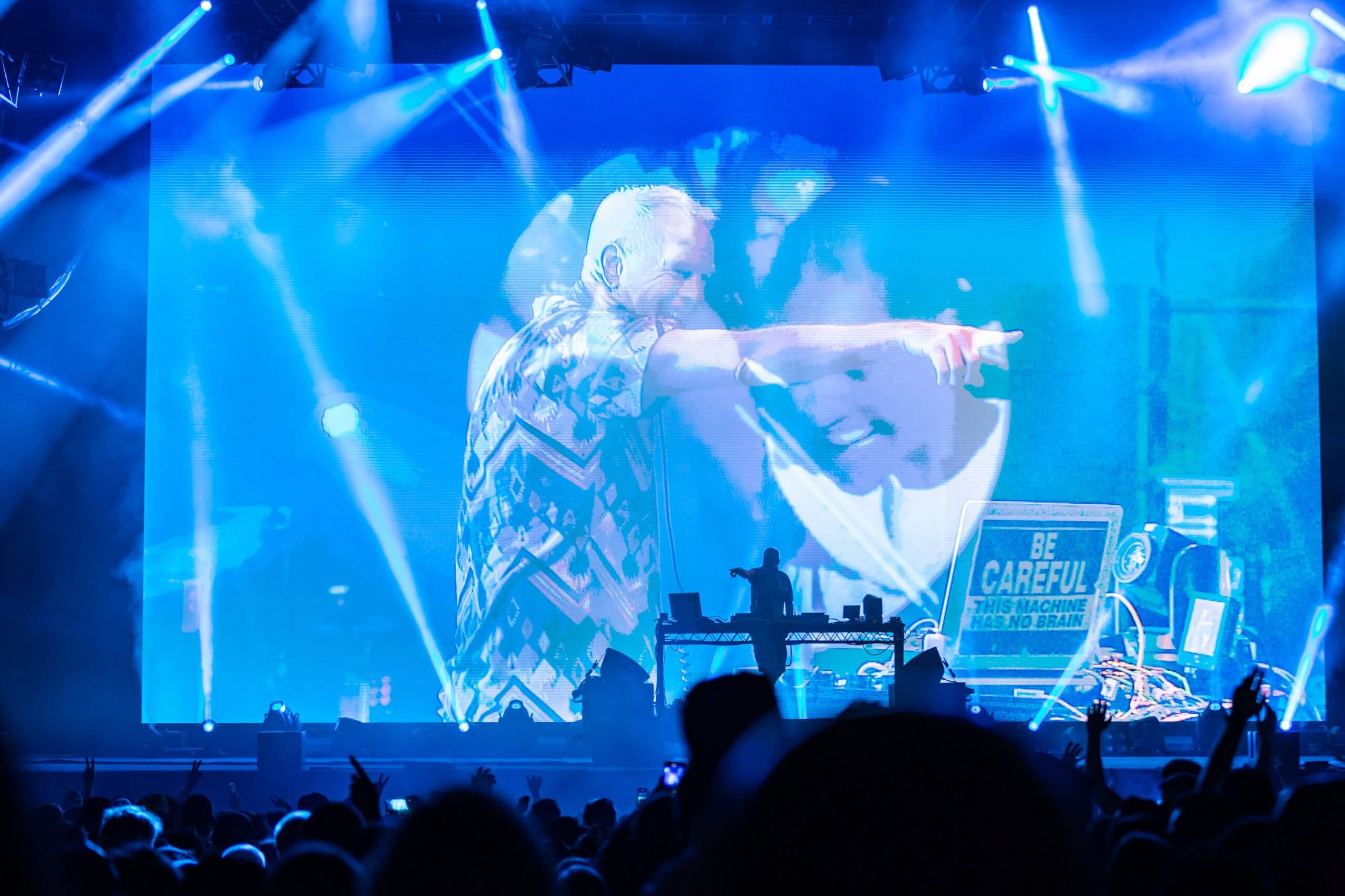 A DJ performs on stage with blue lighting and a large screen behind him displaying his image. The crowd watches as he gestures and points, with bright lights and visual effects creating a lively concert atmosphere.