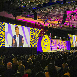 A large audience sits in a dark auditorium watching a speaker on stage. The stage features bright yellow and purple abstract designs with CL23 displayed. The speaker is shown on a big screen behind him.
