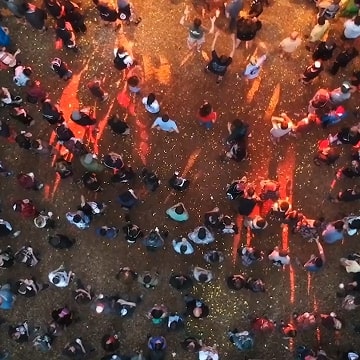 Aerial view of a crowd of people gathered on a reddish-brown ground, with long shadows cast by evening sunlight.