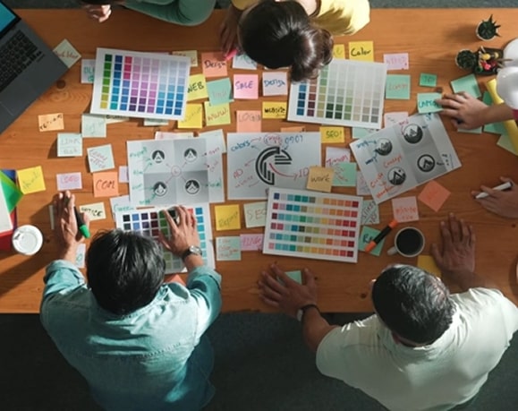 Three people sit around a table covered with color swatches, logo design prints, and sticky notes, collaborating on a creative project. A laptop, coffee cup, and stationery items are also visible.