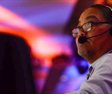 A man wearing glasses and a headset with a microphone looks ahead thoughtfully, with colorful, blurred lights in the background.
