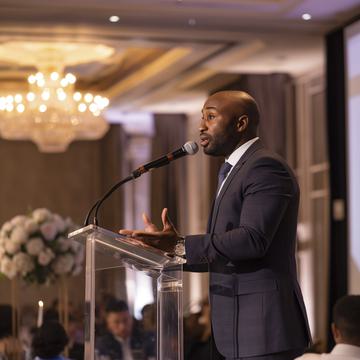 A man in a suit speaks into a microphone at a clear podium during an indoor event, with a chandelier and floral centerpiece visible in the elegant, softly lit room.
