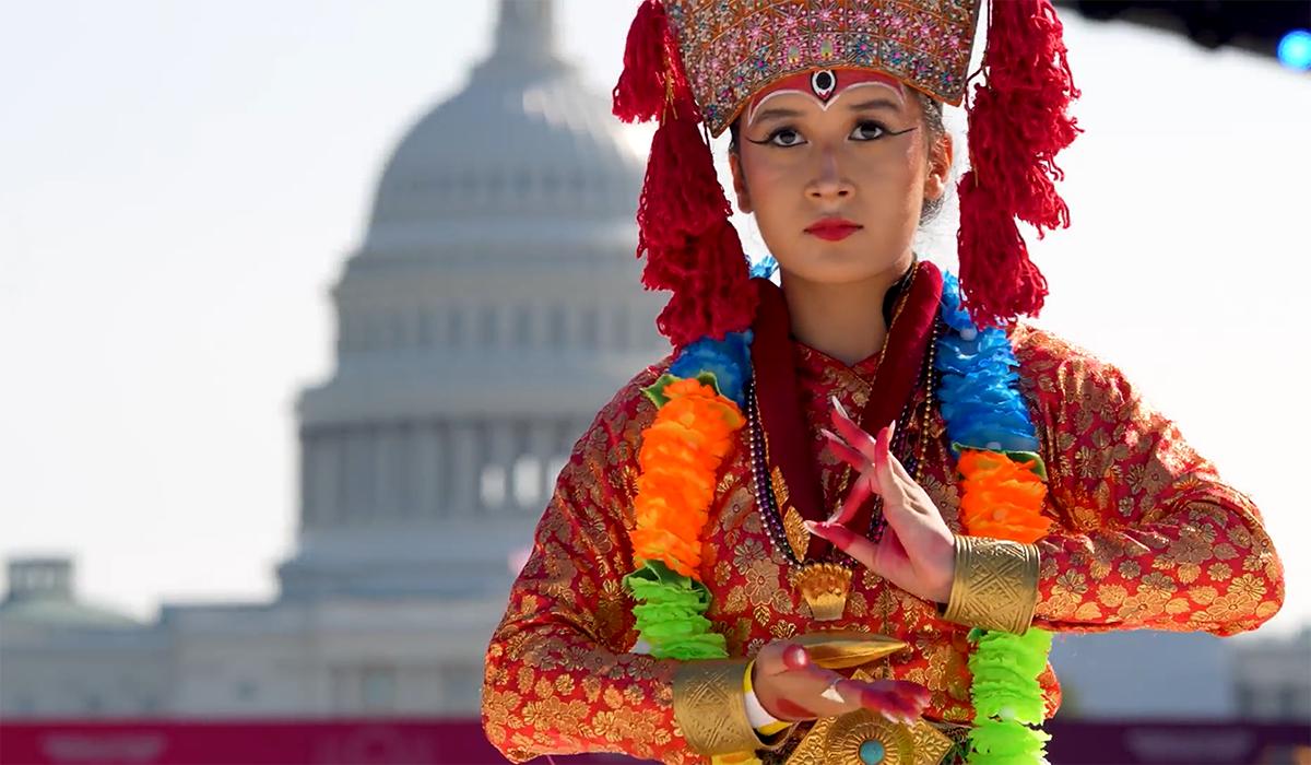 A woman in traditional, colorful Nepali attire and ornate headpiece performs a dance in front of the U.S. Capitol building, with her hands positioned in an intricate gesture.