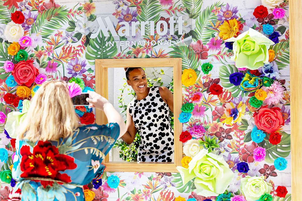 A woman poses for a photo in front of a floral, colorful Marriott International backdrop while another person takes her picture. The backdrop features large paper flowers and tropical leaves.
