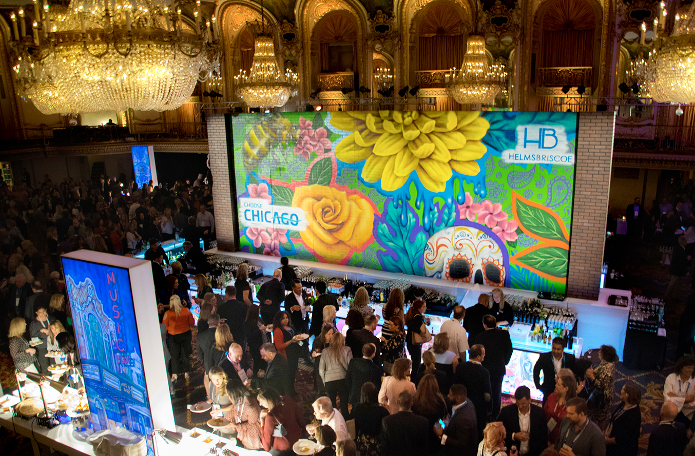 A large crowd gathers in an ornate ballroom with chandeliers, facing a colorful mural featuring flowers and a decorated skull, during an event with food and drinks in Chicago.