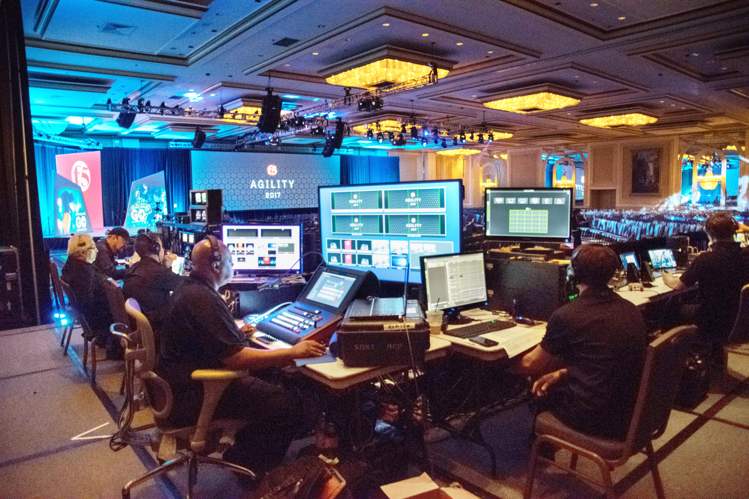 A group of people operate computers and audio-visual equipment in a control area at the back of a large conference room, with multiple screens, stage lights, and an Agility 2019 event banner visible.