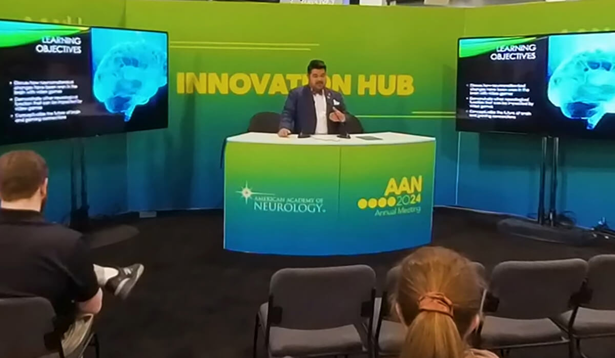 A presenter speaks at the AAN 2024 Annual Meetings Innovation Hub, sitting at a desk with a brain image and learning objectives displayed on two screens. Audience members are seated in front.