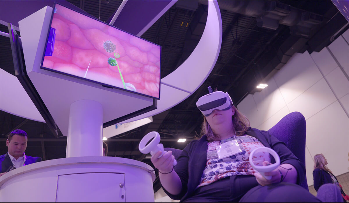 A woman wearing a VR headset and holding controllers sits in a chair, engaged in a virtual reality simulation. A monitor nearby displays a medical-themed VR environment. People and booths are visible in the background.