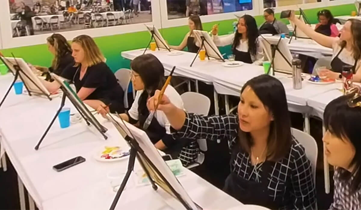 A group of people sit at long tables, painting on canvases with brushes. Art supplies like paint palettes and cups are on the tables. The setting appears to be a classroom or art workshop.