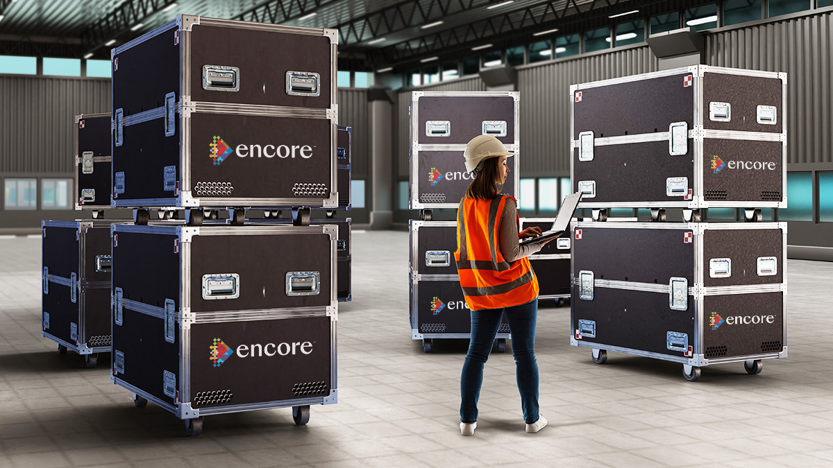 A worker in a hard hat and safety vest uses a laptop in a warehouse with large rolling black equipment cases labeled Encore stacked on the floor around her.