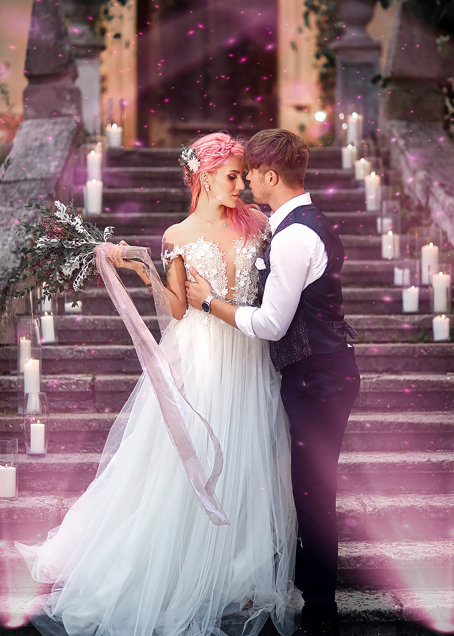 A bride and groom embrace on stone steps decorated with candles. The bride wears a flowing white gown and holds a bouquet with pink ribbons; the groom wears a vest and tie. Pink light and petals float around them, creating a romantic atmosphere.