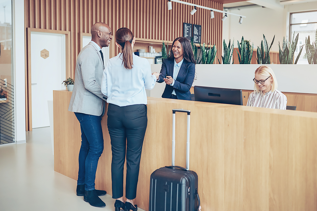 Two people are checking into a hotel at the front desk, speaking with a smiling receptionist. Another woman is seated at a computer. A suitcase is on the floor in front of the counter.