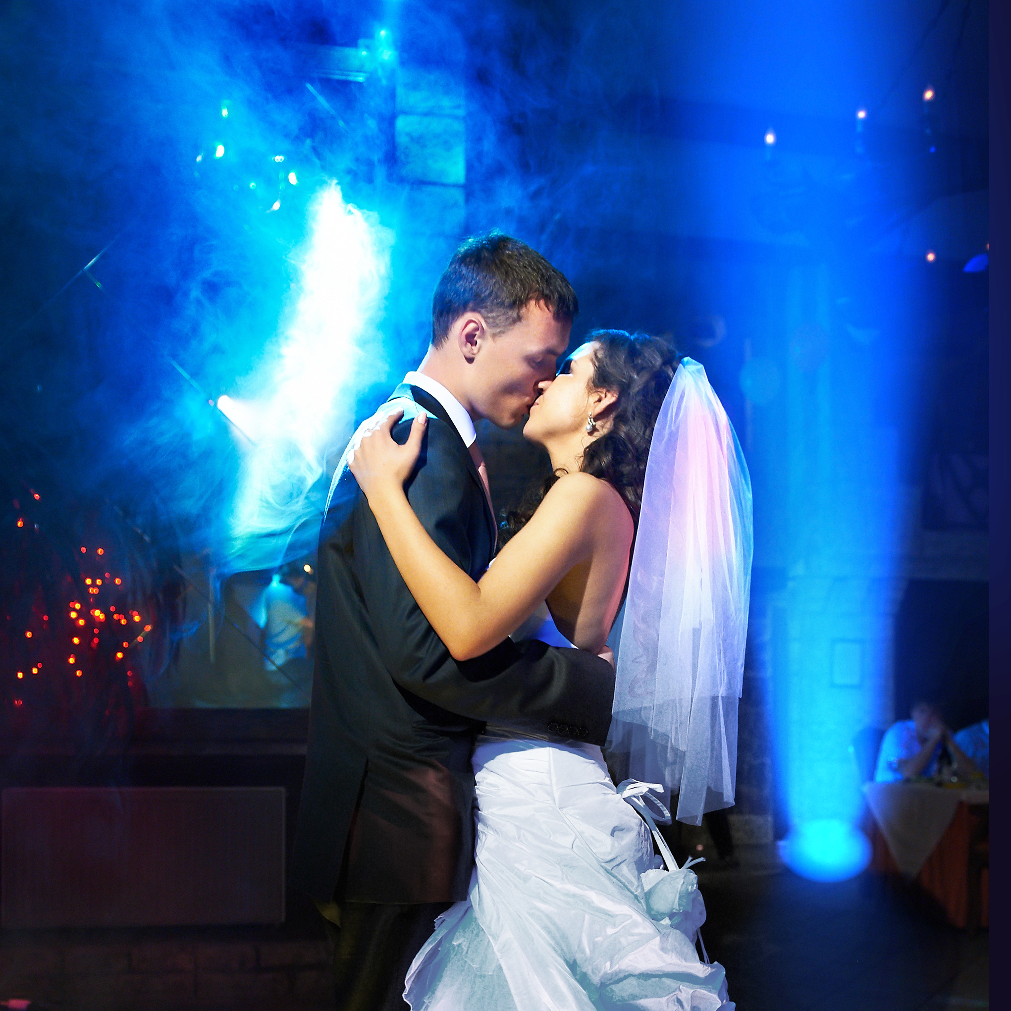 A bride and groom share a romantic dance under blue lights and fog at their wedding reception, wearing formal wedding attire and embracing closely.