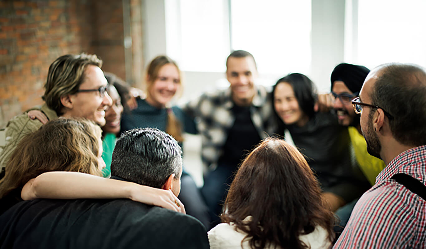 A group of people sit closely together in a circle, smiling and with arms around each other, suggesting a supportive and friendly atmosphere in a bright indoor setting.
