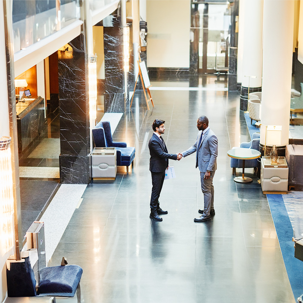 Two men in business attire shake hands in the lobby of a modern building with polished floors, lounge chairs, and large windows letting in natural light.