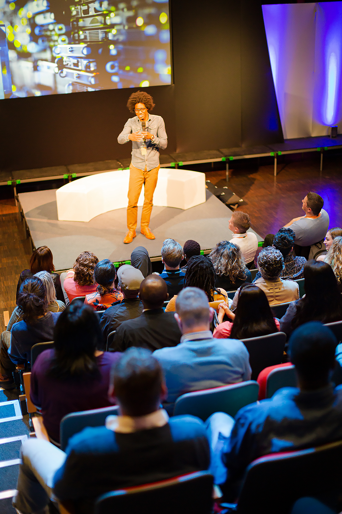 A speaker stands on a stage in front of a large screen and addresses an audience seated in rows in an auditorium. The audience is watching attentively. The atmosphere is professional and engaging.
