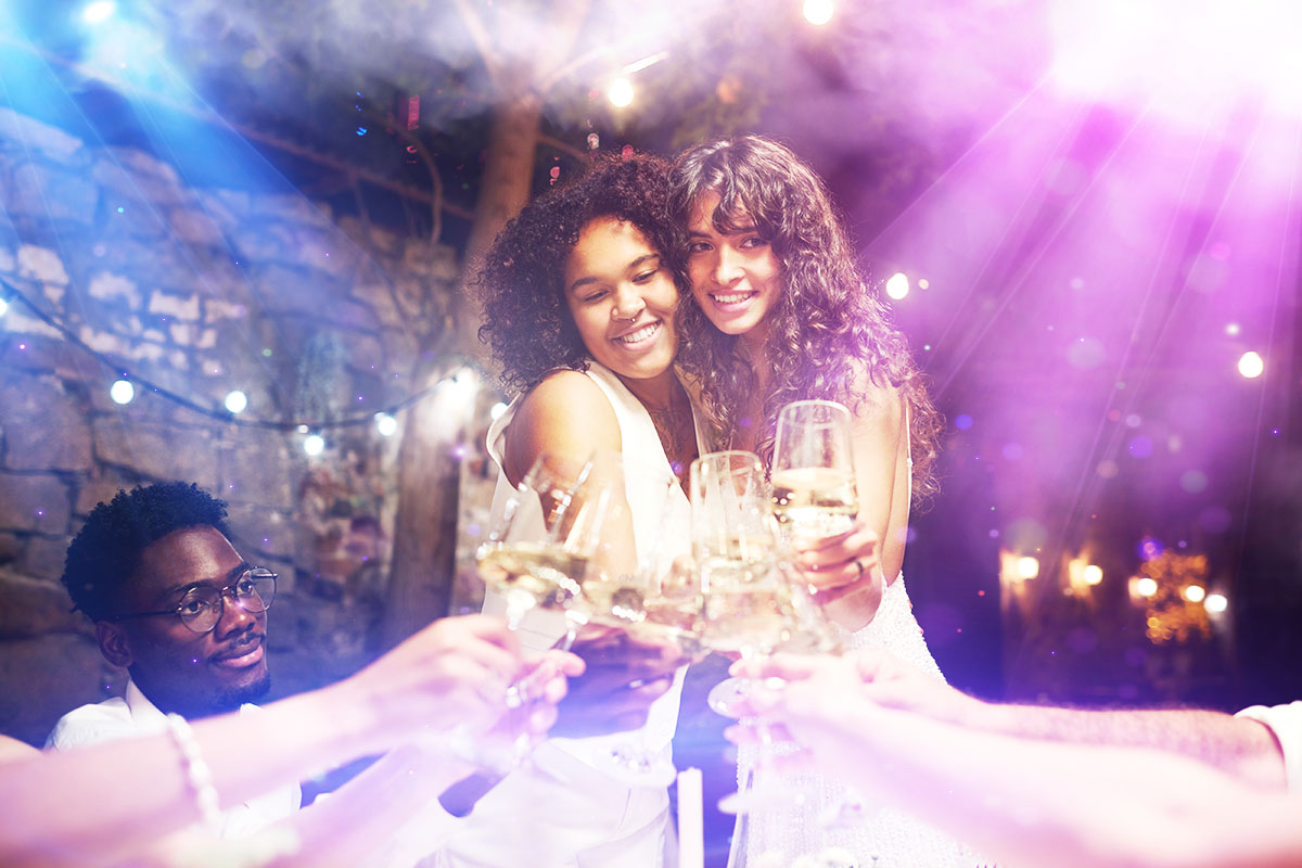 Two women embrace and smile as they toast with friends at an outdoor evening celebration, surrounded by festive lights and colorful party lighting effects.