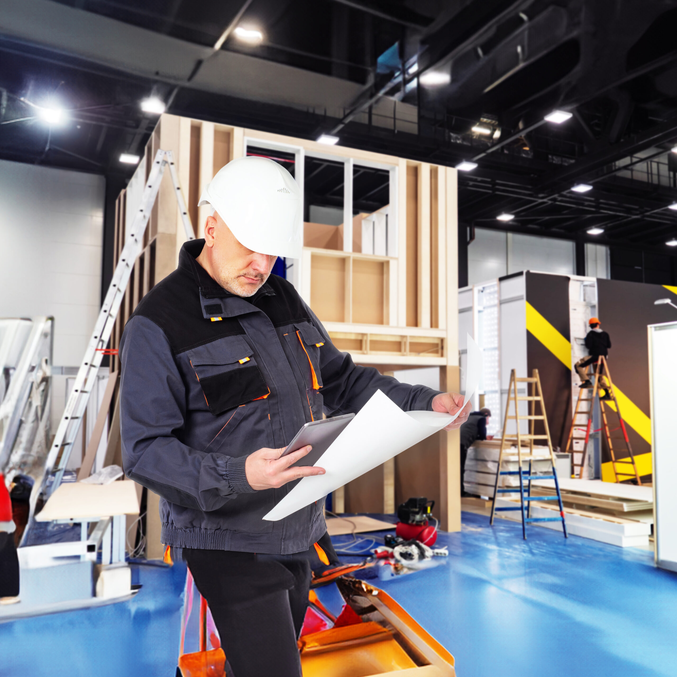A construction worker wearing a white hard hat and dark jacket reviews blueprints inside a modern building under construction, with ladders, tools, and partially finished walls in the background.