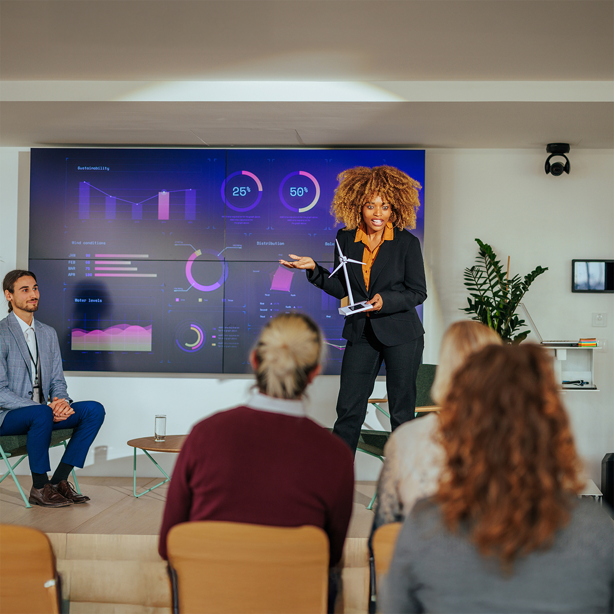 A woman stands presenting data and charts on a large screen to an audience, while a man sits beside her. The audience listens attentively in a modern conference room.